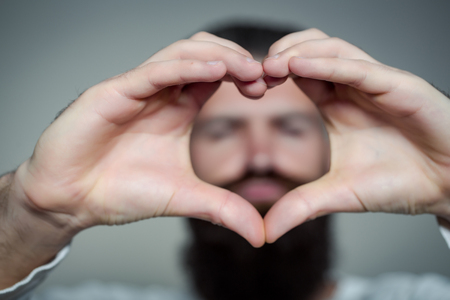 handsome young man with long beard and moustache on face holding hands in heart shape on grey...