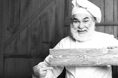 Bearded man cook with smiling face holding and showing on empty wooden plate in chef uniform, black and whiteの写真素材