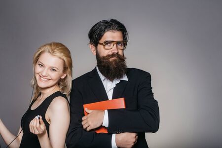 Elegant couple of smiling young woman in black dress listens to music in smartphone with earphones and handsome bearded hipster man in glasses suit keeps folder on grey wallの写真素材