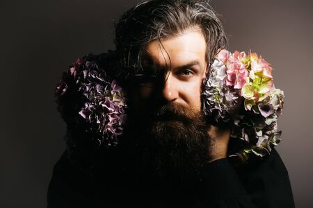 handsome sexy bearded young man hipster with floral beard and mustache on serious face in studio holding hydrangea flower bouquet on grey background, closeupの写真素材