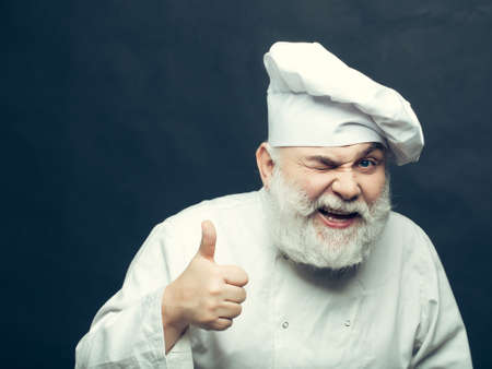 Bearded man cook with winking face in chef hat with thumb up in studio on black backgroundの写真素材