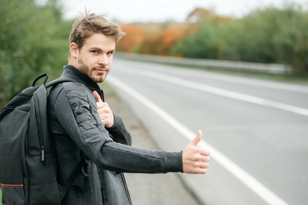Young guy with beard on serious handsome face in casual clothes black backpack standing near road way hitchhiking autostop with hand outdoorの写真素材