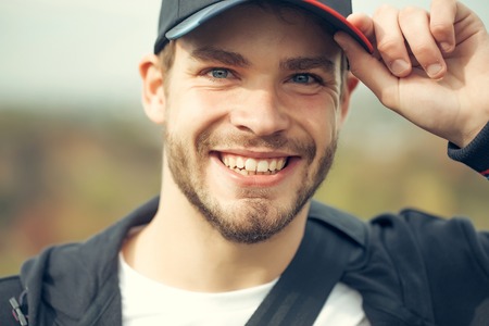 Young guy with beard on his handsome smiling face blue eyes in casual clothes with dark baseball cap on head tipping by hand outdoorの写真素材