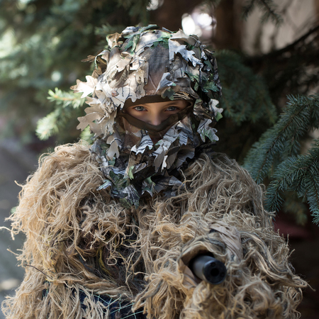 Young girl soldier defender with blue eyes face closed by mask in ghillie camouflage military ammunition with gun standing on background of spruce treeの写真素材