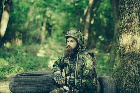 Young soldier wirh bearded dusty tired face in military camouflage uniform with hanging camera and rifle in hand sitting on guard near tree and black tyres in forestの写真素材