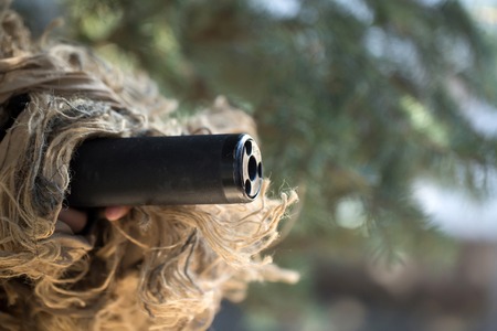 Black muzzle of gun covered with military ghillie mask on green trees in forest background closeup