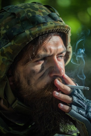 Soldier man with beard on dirty tired face in military camouflage and helmet holding cigarette by hand and smoking outdoor closeupの写真素材