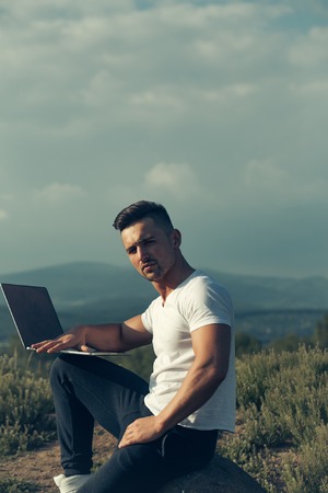Young handsome man with muscular sexy body sitting with laptop outdoor sunny day on blue sky backgroundの写真素材