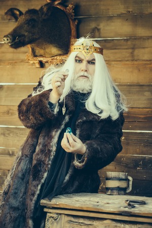 Old bearded man wizard in golden crown with long white hair and beard holding blue gem stone and...