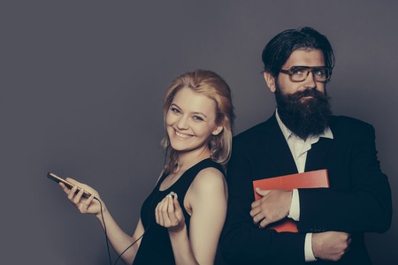 Elegant couple of smiling young woman in black dress listens to music in smartphone with earphones and handsome bearded hipster man in glasses suit keeps folder on grey wallの写真素材