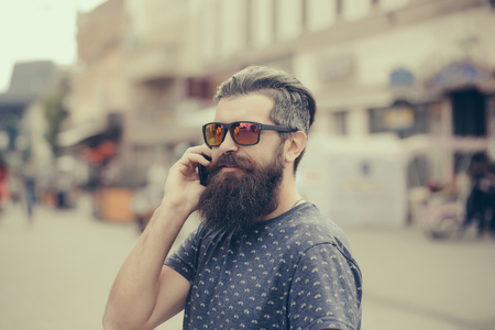 handsome bearded young man hipster with long beard and mustache has stylish hair on smiling hairy face in sun glasses speaking on mobile phone outdoor, closeupの写真素材