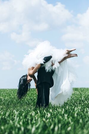 young wedding couple of girl with brunette hair and pretty face in white bride dress and handsome man in black groom suit on green grass in field on cloudy blue sky natural backgroundの写真素材