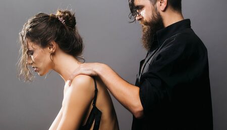 young couple of pretty woman in black jeans and bra has slim body and handsome bearded man with long beard in shirt doing massage on grey backgroundの写真素材