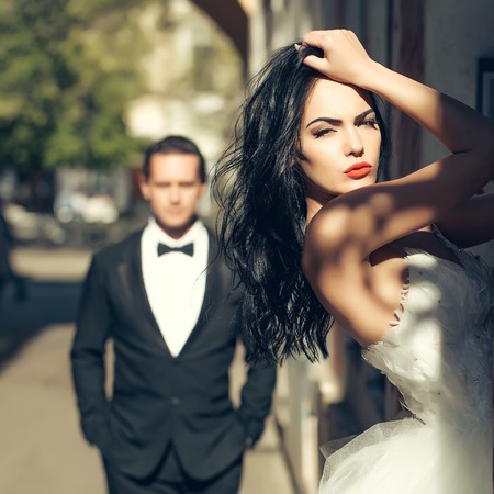 young wedding couple of girl with brunette hair and red lips on pretty face in white bride dress and handsome man in black groom suit standing near building arch sunny outdoorの写真素材
