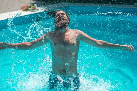 Young man with smiling bearded face wet hair and body splashing water and having fun in summer pool with blue waterの写真素材