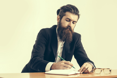 young handsome bearded man scientist or professor with long beard and teacher glasses with pencil and book or notepaper sitting at table on light backgroundの写真素材