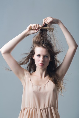 young woman with slim sexy body pretty face and brunette long hair in dress holding hairbrush standing in studio on grey backgroundの写真素材