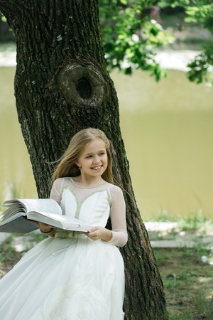 small girl kid with long blonde hair and pretty smiling happy face in prom princess white dress standing sunny day outdoor near water with bookの写真素材
