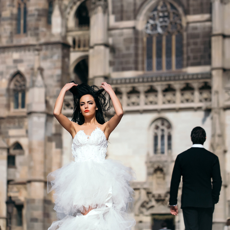 young wedding couple of girl with brunette hair and red lips on pretty face in white bride dress and handsome man in black groom suit near castle or palace building sunny outdoorの写真素材