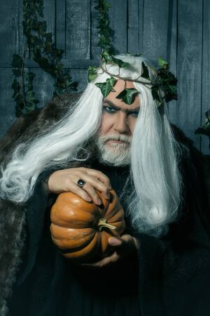 Old grey-haired man in costume of evil sorcerer with wreath of ivy and pumpkin on Halloweenの写真素材