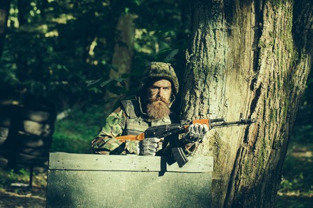 Young soldier hipster with beard on dirty tired face in military ammunition and helmet standing on guard near tree and wooden board with gun in hands in forestの写真素材