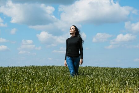 young girl or woman with brunette hair and pretty face in black cardigan and jeans on green grass in field on cloudy blue sky natural backgroundの写真素材