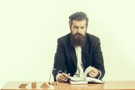 young handsome bearded man scientist or professor with long beard and teacher glasses with book or notepaper sitting at table with wooden cubes isolated on white backgroundの写真素材