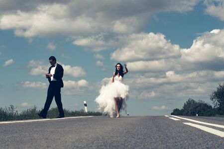 young wedding happy couple of girl with brunette hair and pretty face in white bride dress and handsome man in black groom suit on road way on cloudy blue sky nature backgroundの写真素材