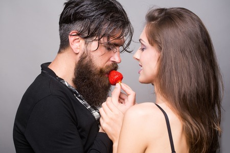 young sexy couple of woman with pretty smiling face and brunette hair with bare shoulders and handsome bearded man with long beard eating red strawberry berry fruit in studio on grey backgroundの写真素材