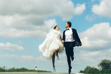 young wedding happy couple of girl with brunette hair and pretty face in white bride dress and handsome man in black groom suit running on road way on cloudy blue sky nature backgroundの写真素材