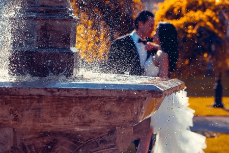 young wedding couple of sexy pretty girl or woman with hair in white bride dress and handsome man in black groom suit kiss near fountain with splashes of water drops sunny outdoor with yellow treesの写真素材