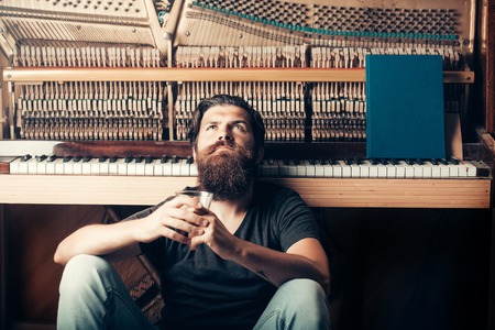 handsome bearded man with stylish hair mustache and beard on serious face drinking brandy or whiskey from glass near old wooden open piano with keyboard as musician with music book, copy spaceの写真素材