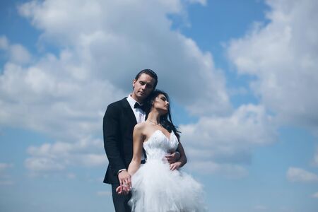 young wedding couple of sexy girl with brunette hair and pretty face in white bride dress and handsome man in black groom suit on cloudy blue sky natural backgroundの写真素材