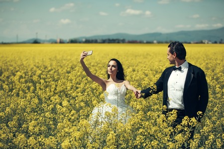 young wedding couple of sexy girl with pretty face in white bride dress holds mobile phone and handsome man in black groom suit in field with yellow flowers on natural background with blue skyの写真素材