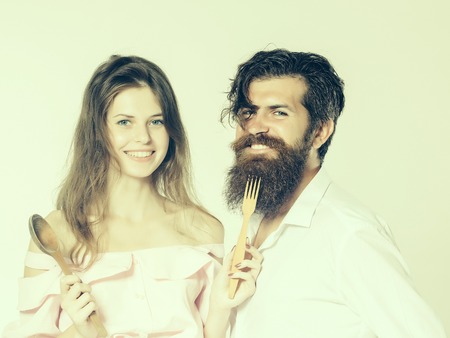young smiling happy couple of pretty girl with long hair and handsome bearded man with beard holding wooden spoon and fork isolated on whiteの写真素材