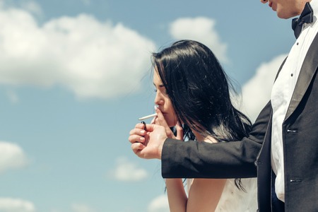 Man hand offers to woman beautiful brunette gas lighter to light cigarette for smoking on blue skyの写真素材