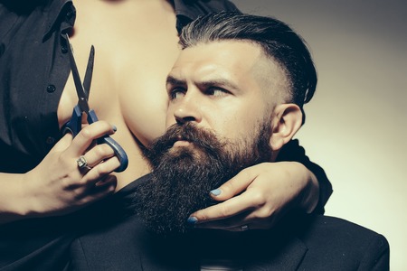 Young handsome man with long beard and moustache on serious face with female beautiful bare chest of young woman cutting with scissorsの写真素材