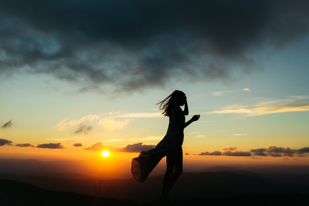 woman or girl silhouette walking at beautiful view on sunset or sunrise in blue sky with clouds and yellow orange sun in mountains on natural backgroundの写真素材
