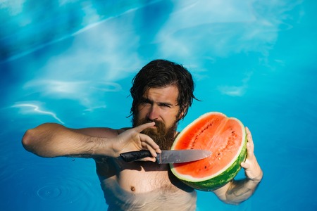young bearded man with wet hair and beard swimming in pool with blue water holds knife and green red watermelon sunny summer day outdoorの写真素材