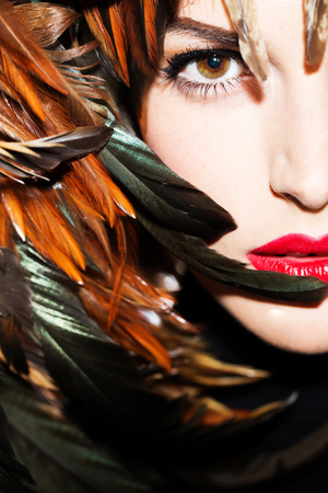 young woman or girl with red lips on pretty face in beautiful feather hat brown color as halloween or carnival holiday costume, closeupの写真素材