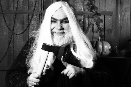 Old man witch in robe with demonic grin hold hatchet near finger in wooden house, black and whiteの写真素材