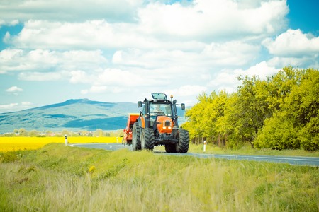 Red farm tractor drives on road along beautiful field on summer sunny day on landscape backgroundの写真素材