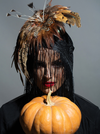 young woman or girl with red lips on pretty face in beautiful feather hat brown color as halloween or carnival holiday costume and veil holds pumpkin on grey backgroundの写真素材