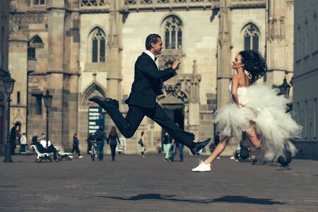 young wedding happy couple of girl with brunette hair and pretty face in white bride dress and handsome man in black groom suit jumping near castle or palace building sunny outdoorの写真素材
