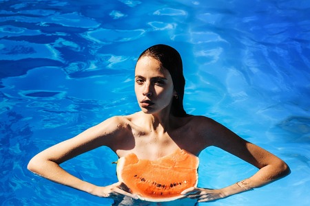 young woman or girl with pretty face and wet hair swimming in pool with blue water eating red watermelon sunny summer day outdoorの写真素材