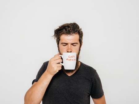 handsome bearded man with stylish hair beard and mustache on serious face in shirt holding white cup or mug with good morning text drinking tea or coffee in studio on grey backgroundの写真素材