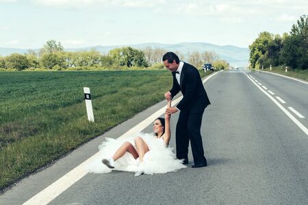 young wedding happy couple of girl with brunette hair and pretty face in white bride dress and handsome man in black groom suit on road way on cloudy blue sky nature backgroundの写真素材