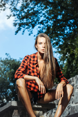 pretty slim model girl with long brunette hair in red checkered shirt and shorts sits on stone fenceの写真素材