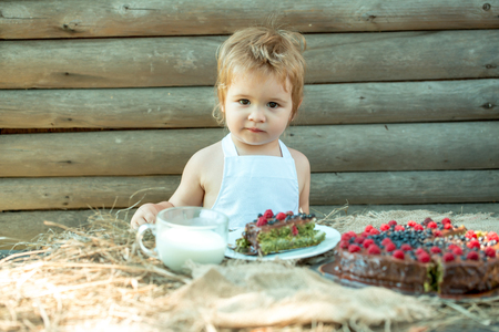Cute little boy in white pinafore sits at table served with fruit cake and cup of milk outdoors on wooden backgroundの写真素材