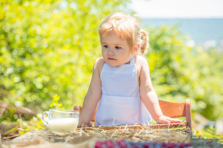 Cute little boy in white pinafore sits in high chair at table with cup of milk on sunny summer day on natural backgroundの写真素材